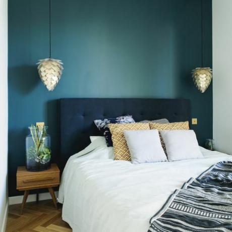 Intérieur de chambre à coucher élégant avec petite table de nuit en bois, jardin dans un pot, literie blanche, coussins de couleurs et couverture. Espace aux murs bleus et parquet en bois marron. Lampe design.