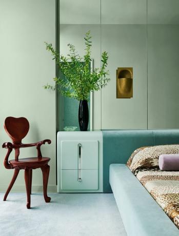 une chambre a des murs vert menthe, un mur en miroir derrière un lit recouvert d'un velours aqua clair et une table de chevet vert clair, une applique en bronze, un fauteuil en chêne avec un dossier en forme de cœur, un tapis vert menthe