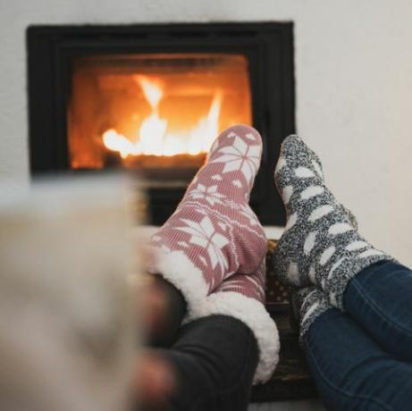 vue de derrière de deux copines assises près de la cheminée et de l'arbre de noël avec leurs pieds avec des chaussettes d'hiver sur une table basse, buvant du café ou du thé