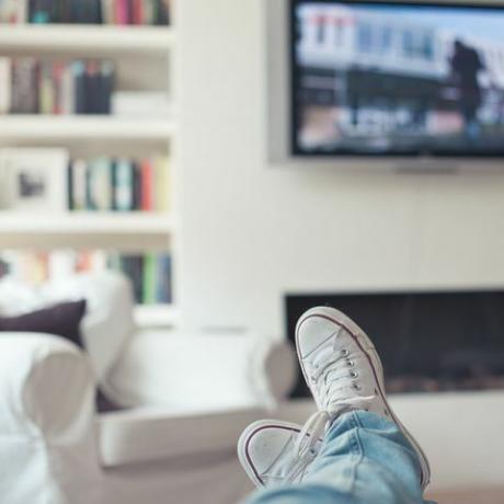 Photo de jeans et de baskets, les pieds sur le canapé, avec télévision, cheminée, canapé et bibliothèque en arrière-plan.